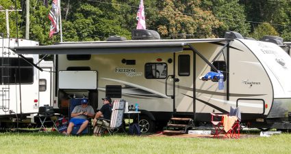 Two fans relax in folding chairs beneath the awning of a parked travel trailer in the campground, surrounded by RVs, camping gear, and NASCAR-themed flags on a sunny day.