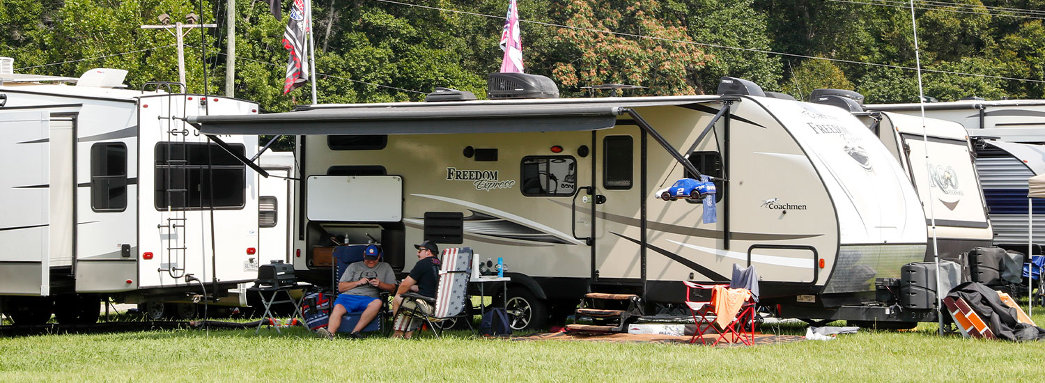 Two fans relax in folding chairs beneath the awning of a parked travel trailer in the campground, surrounded by RVs, camping gear, and NASCAR-themed flags on a sunny day.